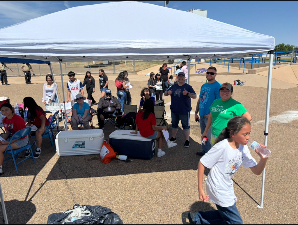 Group of people under a white canopy, some seated and others standing; some are walking or running; several coolers and a bag nearby.