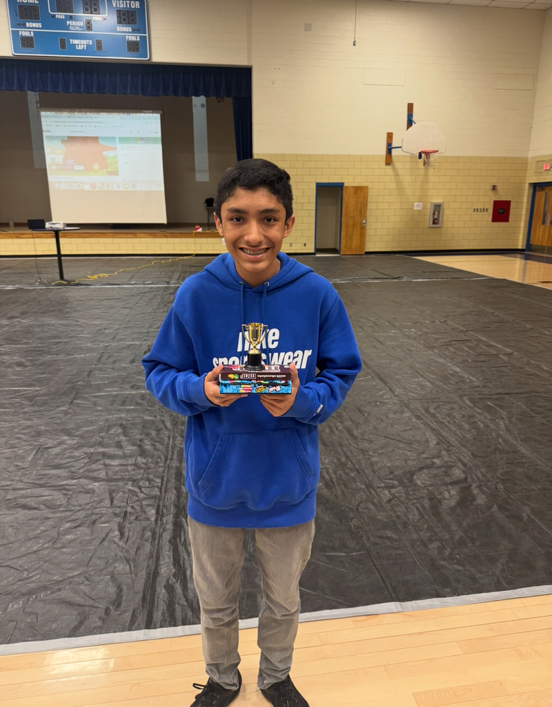 Boy with hoodie, holding a trophy, smiling, standing in an empty gymnasium with a black floor.