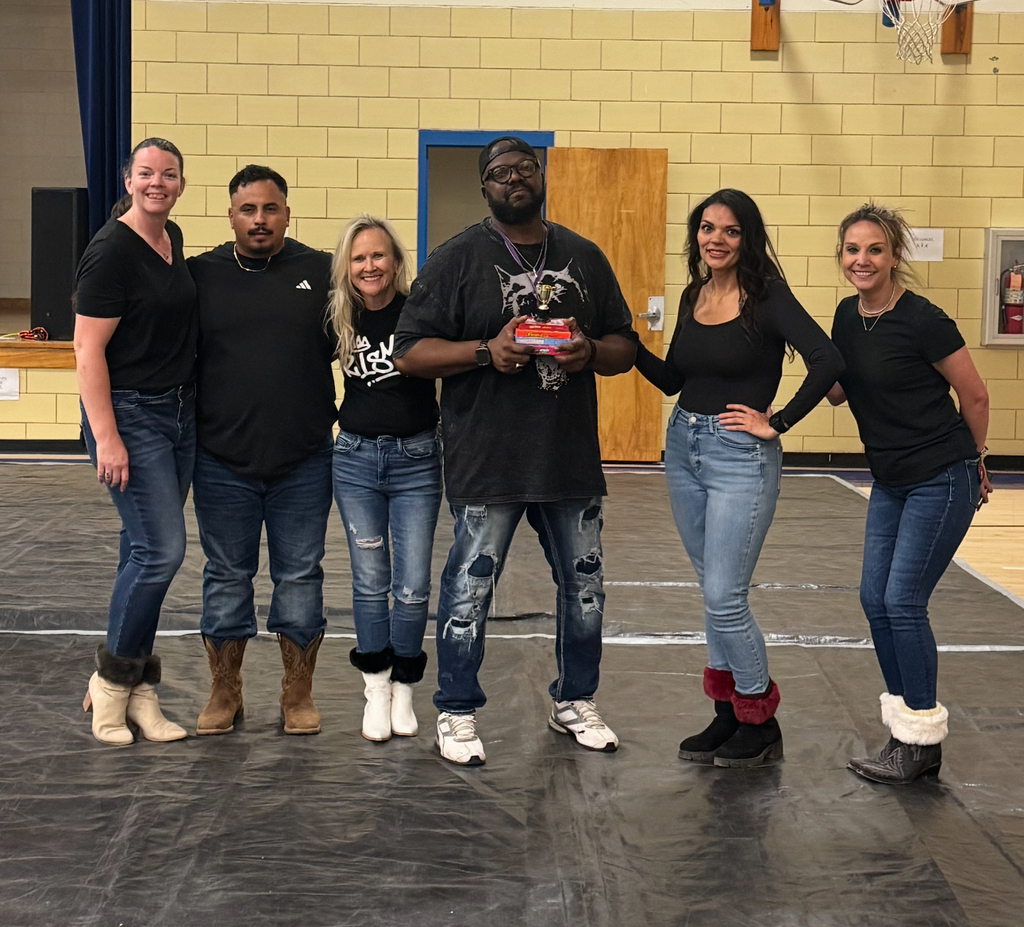 Six individuals, including four women and two men, stand together on a stage in a gymnasium. They wear black shirts and jeans.