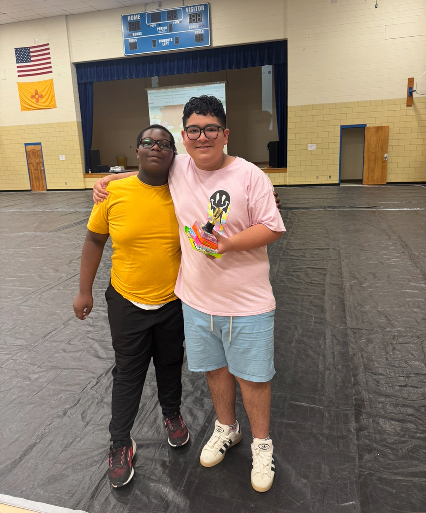 Two individuals stand in a gymnasium with one holding a trophy. One wears a yellow shirt, and the other wears a pink shirt and shorts.