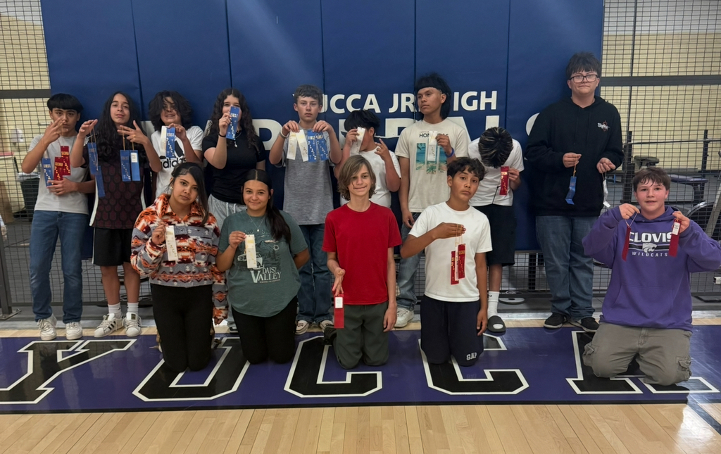 Students in a gymnasium hold certificates. A blue banner reads "Yucca Jr. High." The floor has a purple mat.