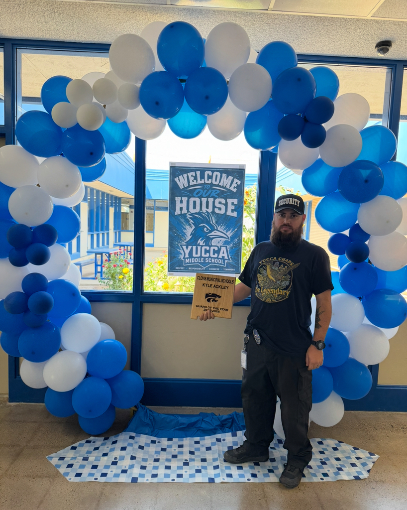 Man with a box standing before a blue and white balloon arch, in front of a school banner.