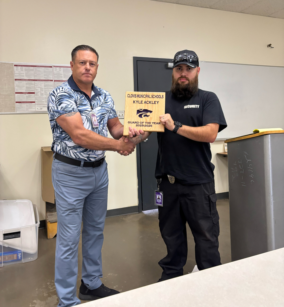 Two men stand indoors; one hands the other a plaque. Behind them is a whiteboard and a door.
