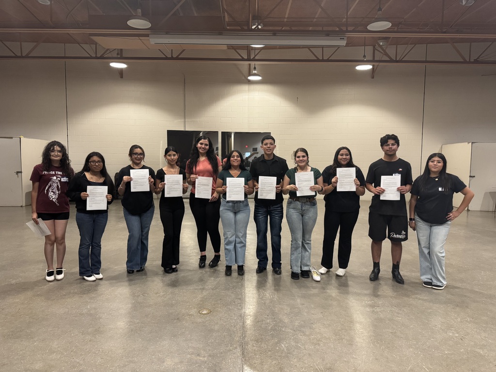 A group of individuals stands in a room holding white papers, possibly certificates. They are dressed casually, with sneakers and jeans, against a background of plain walls.