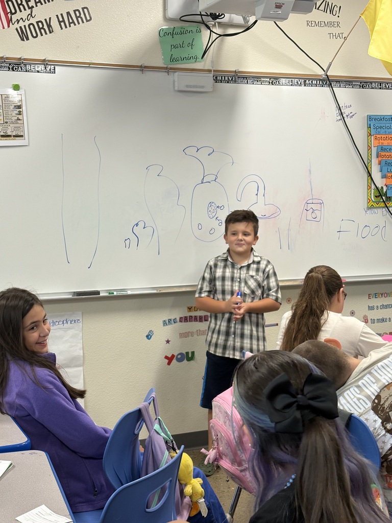 A boy stands at a whiteboard in a classroom, while several students sit in chairs, facing him.