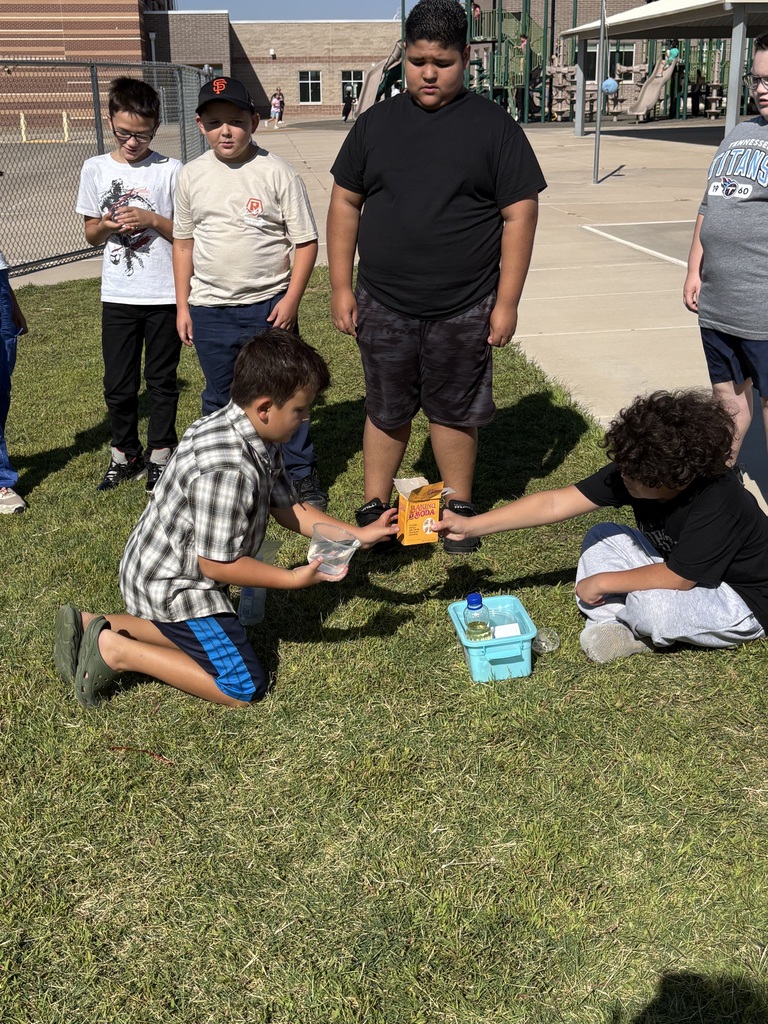 A group of kids in a grassy area, with one kneeling and another standing. They are engaging in an activity.