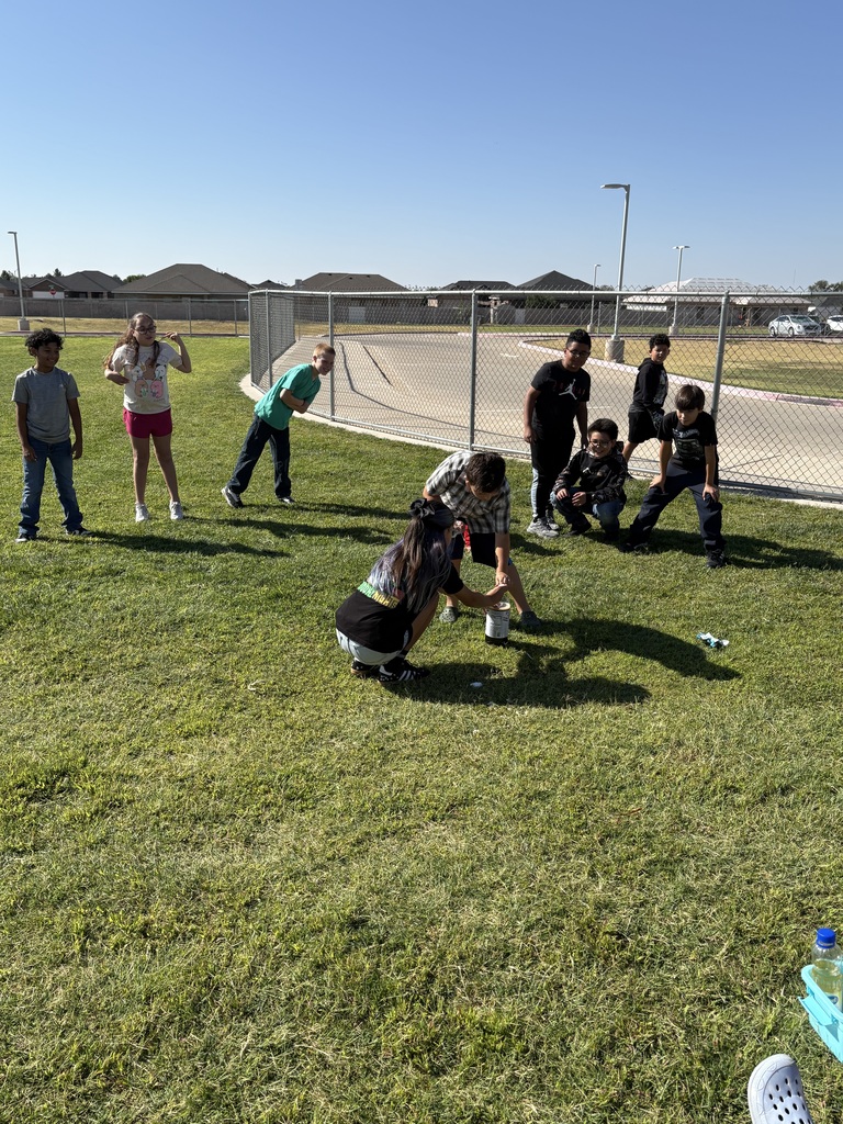 A group of children and adults play on a grassy field, with a chain-link fence in the background.