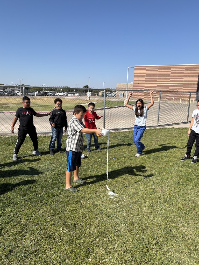 Seven children play on a grassy field with a building in the background. They hold a long white string.