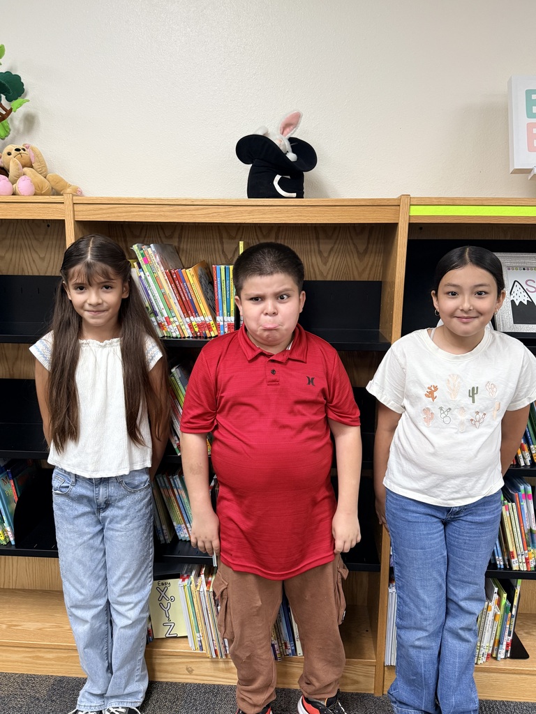 Three children stand together in front of a bookshelf filled with books. They are wearing casual clothing.