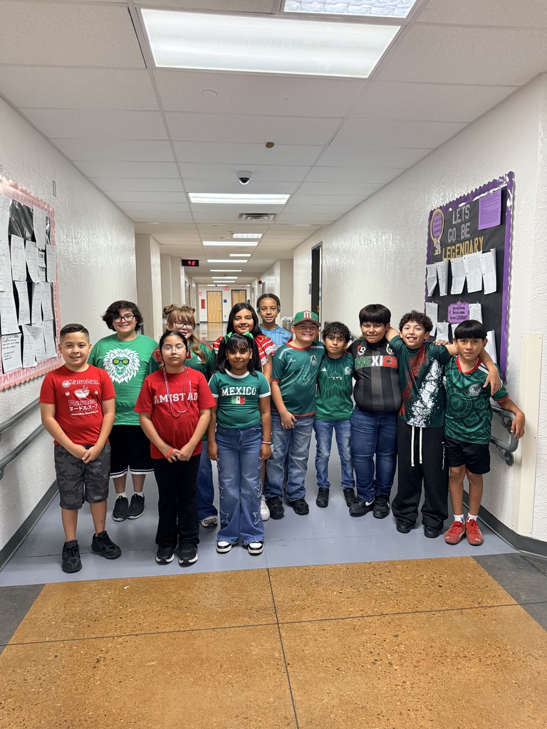 A group of young people wearing green shirts and jeans stand in a hallway with white walls.