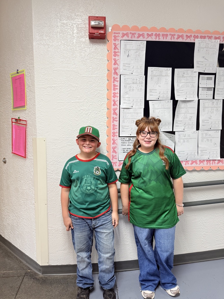 Two children stand together, both wearing green shirts and jeans, in front of a bulletin board with papers.