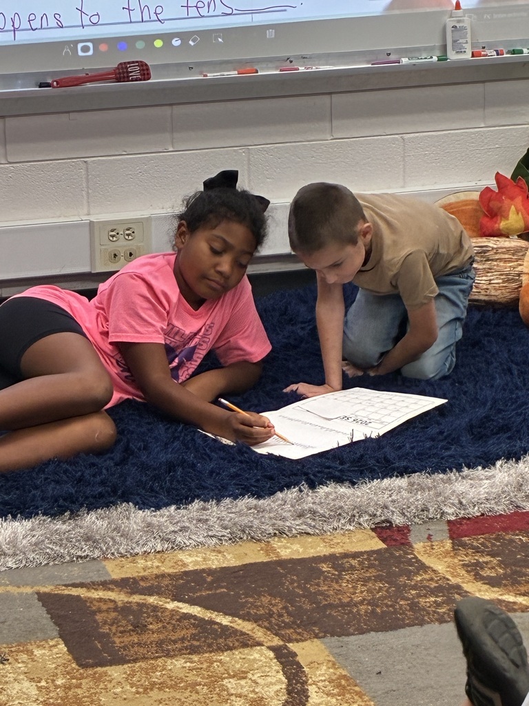Two children on a blue carpet with papers, one writing and the other leaning over a paper.