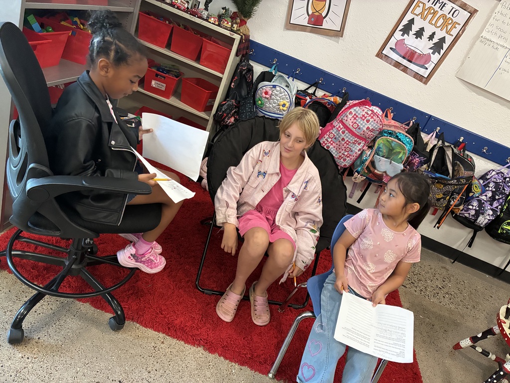 Three children seated on chairs in a classroom with red carpet. One reads a book, another holds paper. Backpacks and posters adorn the room.