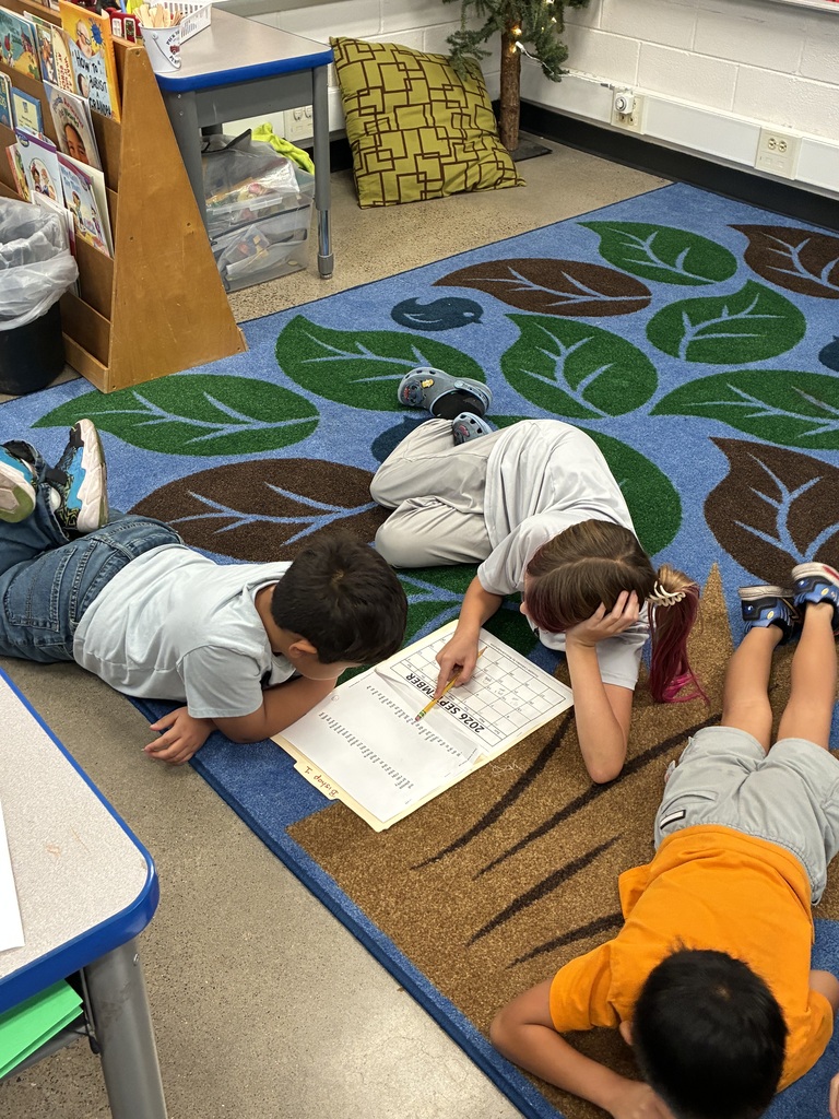 Four young children lie on a rug with leaf designs, working on papers. A small tree stands in the corner.