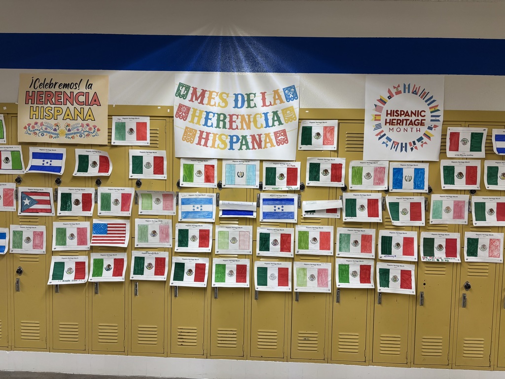 A locker wall with flags from various countries, including the USA, Mexico, and others. Above the lockers, banners with the text "Herencia y Herencia Hispana" and "Celebrate Hispanic Heritage Month" are displayed.