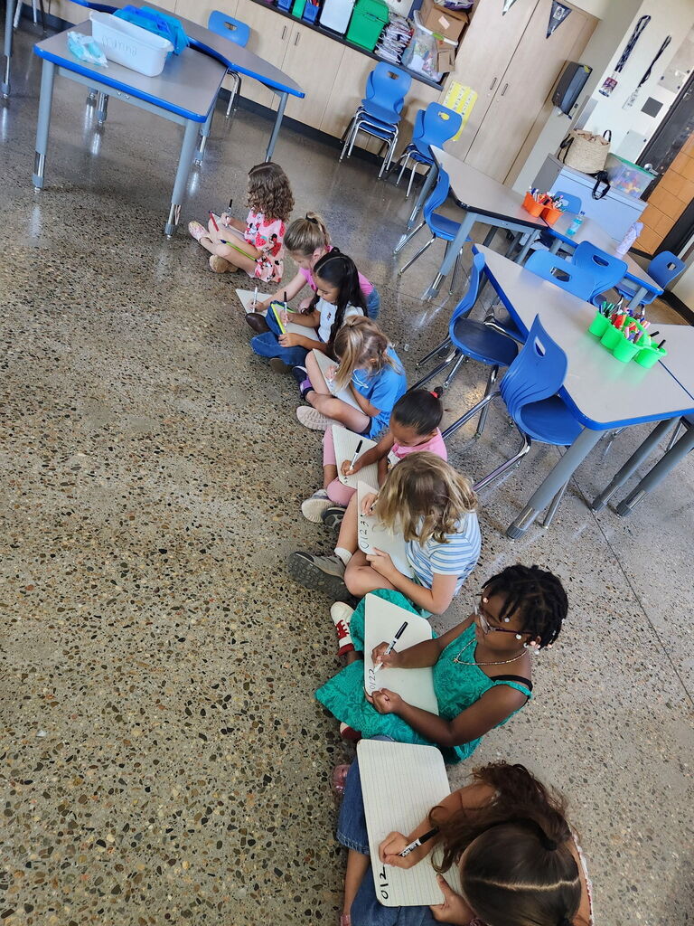 Children seated in a classroom, working on assignments. A teacher at the end of the row.