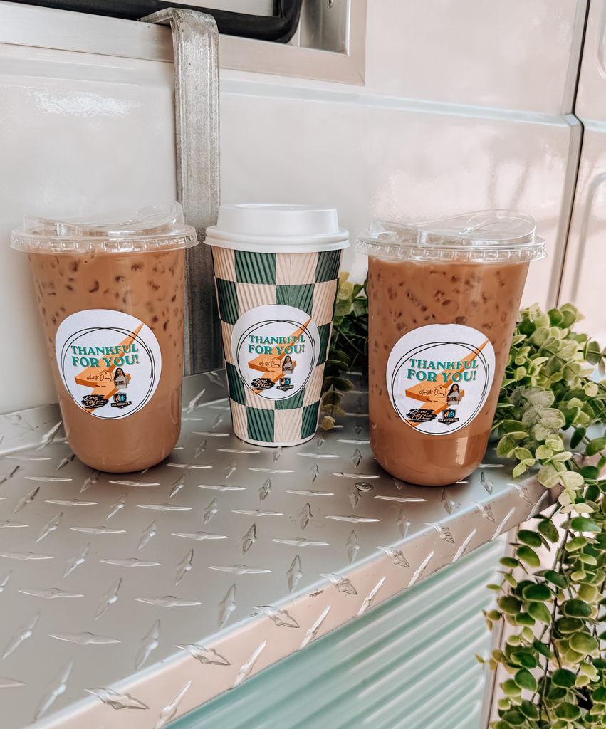 Three iced coffee drinks in plastic cups with white lids on a metal surface with green plants.