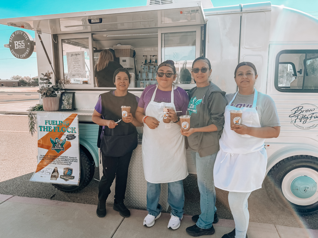 Four women stand by a coffee truck. They hold cups of coffee. A banner hangs beside the truck.