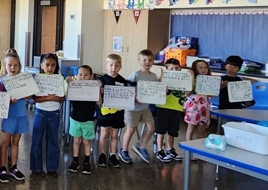 A group of children standing in a classroom, holding whiteboards with writing on them.
