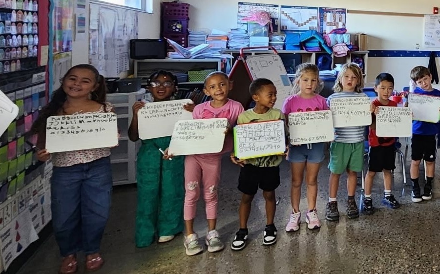 Children in a classroom holding up white signs. Background includes shelves and a bulletin board.