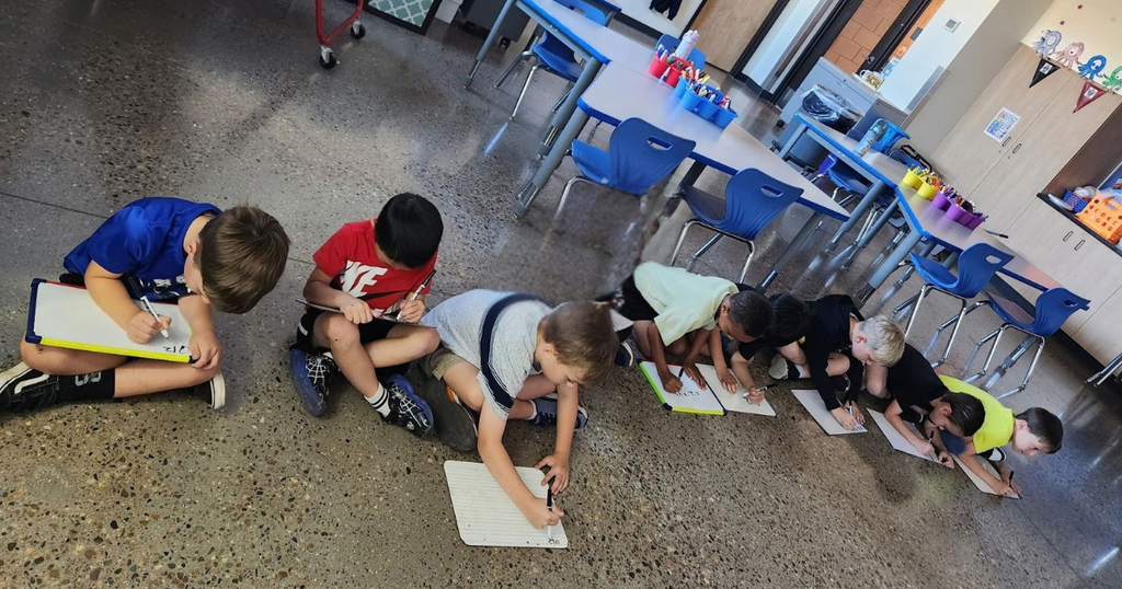 Several children sit or kneel on the floor, each holding a clipboard and pen. Behind them are tables and chairs.