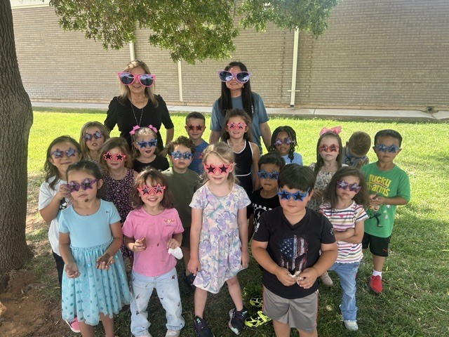 Group of children and adults wearing sunglasses pose under a tree. The background shows a building.