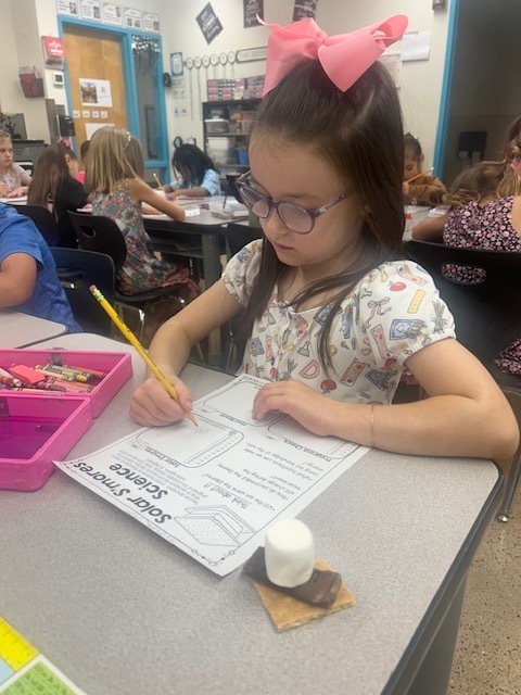 Child in glasses with a pink bow sits at desk, writing on paper with a pencil.