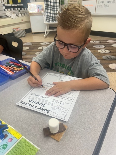 A young boy in glasses sits at a desk, focused on writing on a sheet of paper.