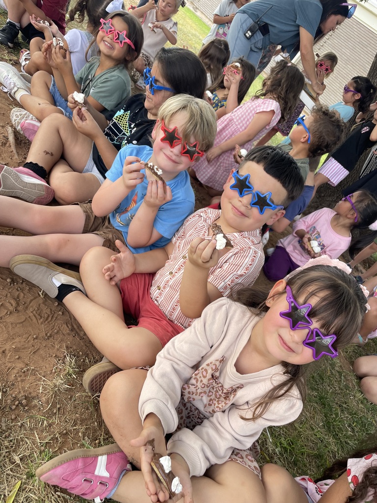 A group of children are sitting on grass wearing patriotic glasses. They are holding snacks. Other people sit behind them.