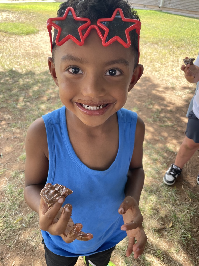 A child with brown hair in a blue tank top, wearing star-shaped headband, holds a chocolate bar.