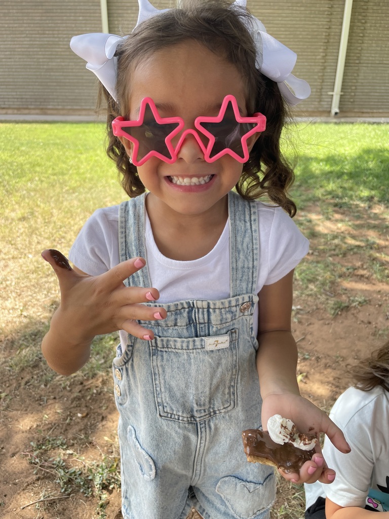 A child in white sunglasses, a white shirt, and denim overalls holding a dessert outdoors.
