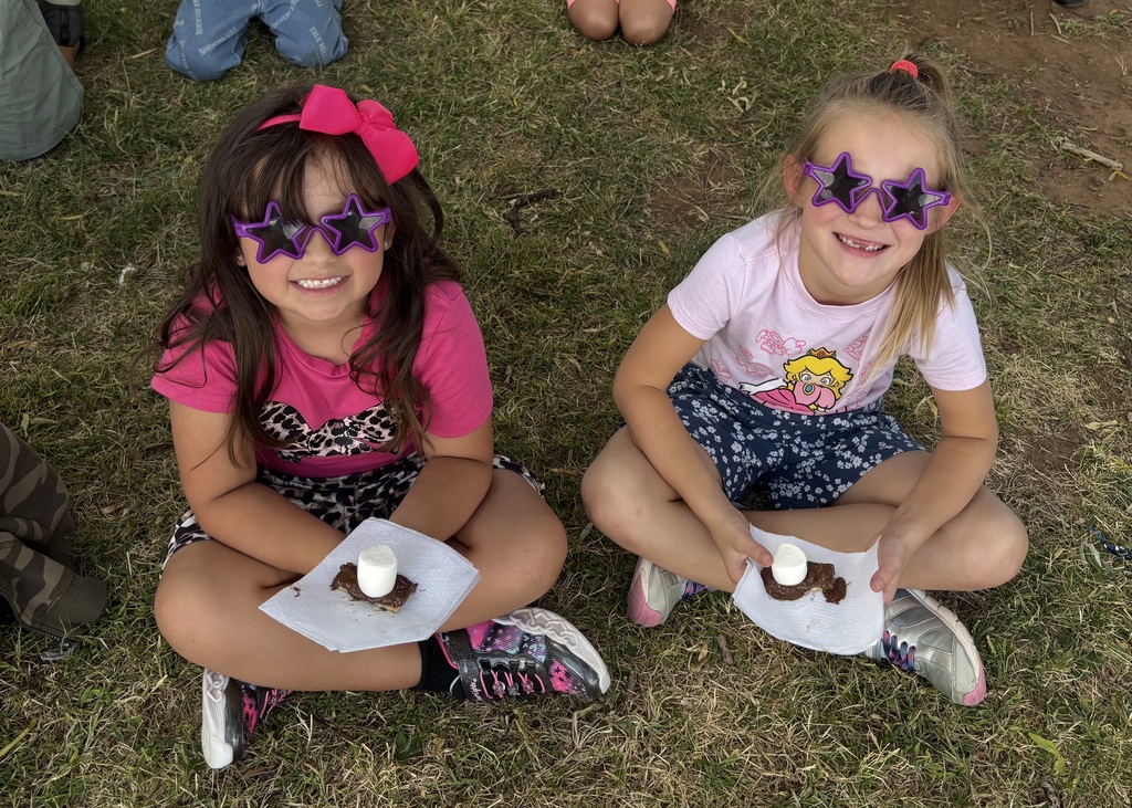 Two young girls sit on grass with desserts. Each holds a plate with a treat and wears sunglasses.