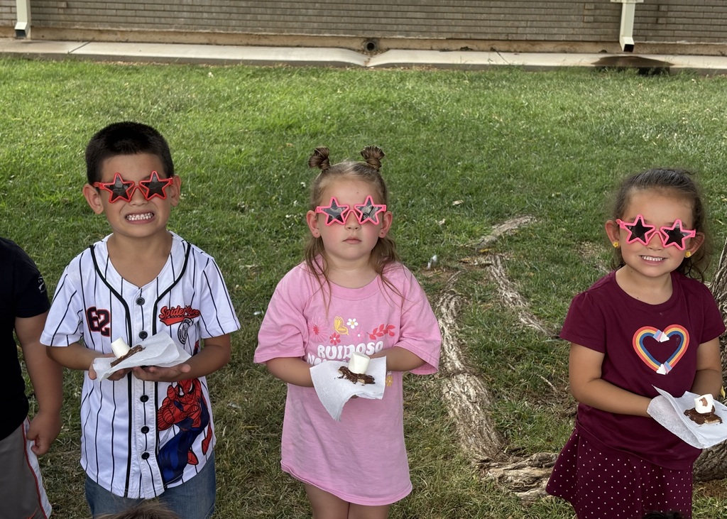 Four children, wearing star-shaped glasses, stand on grass holding food. A building is in the background.