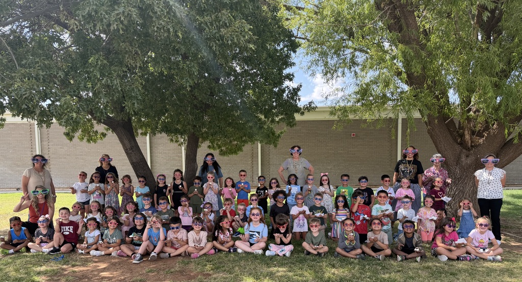 Group of children and adults gathered outdoors in a park, some sitting on grass, others standing, under trees.