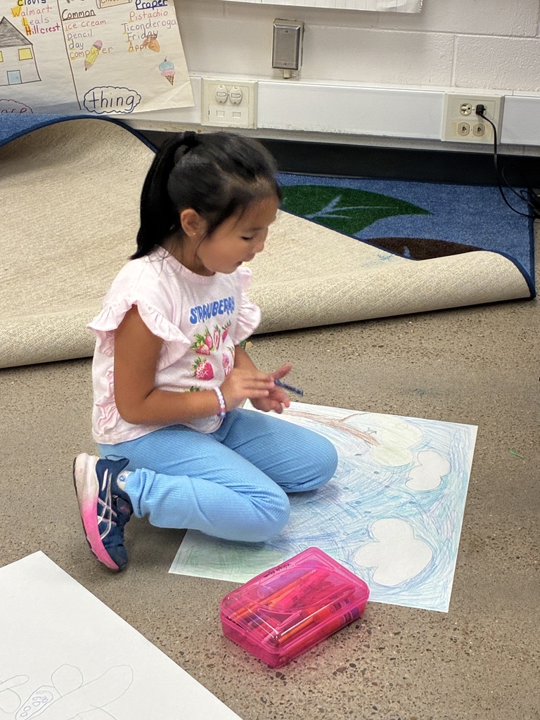 A young child wearing a pink top and blue pants is seated on the floor, drawing on paper.