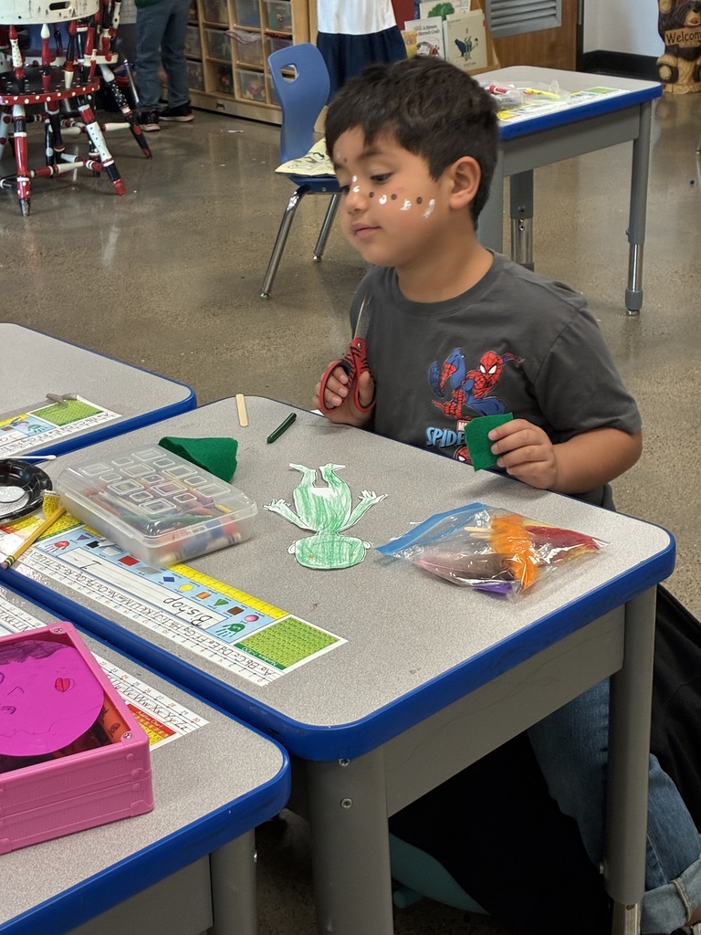 A boy at a desk with a green toy and a green cup. Other tables and chairs are in the background.