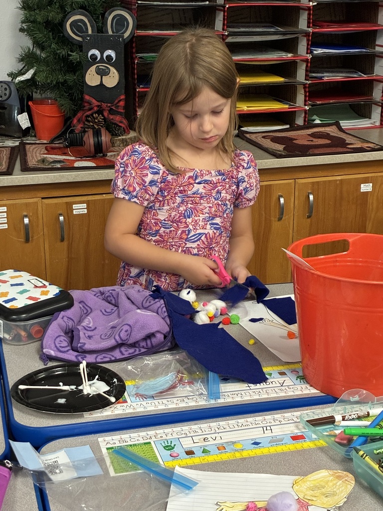 A young girl is cutting fabric with scissors in a classroom. The desk has art supplies.