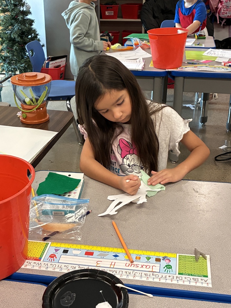 A young girl seated at a table is working on a craft project. She has long hair and is surrounded by various art supplies.