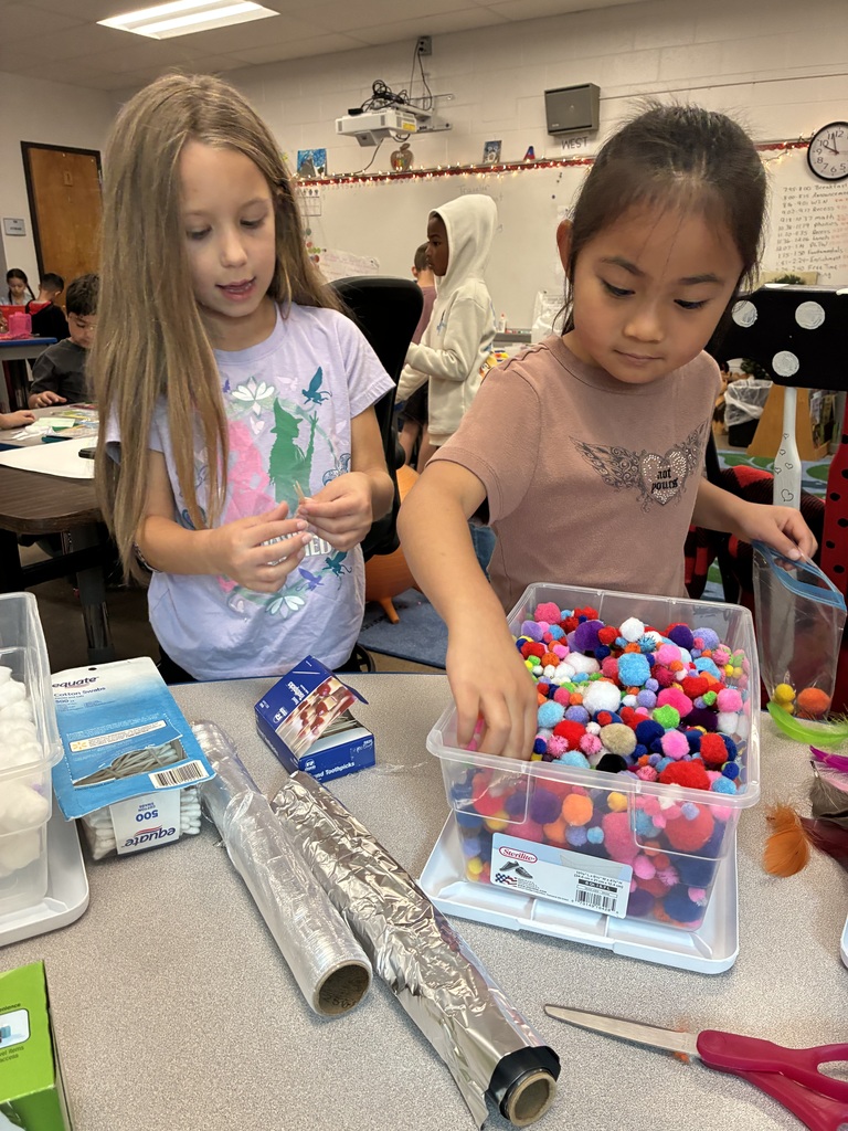 Two young girls working on a project in a classroom. One is reaching into a bin of colorful pom poms.