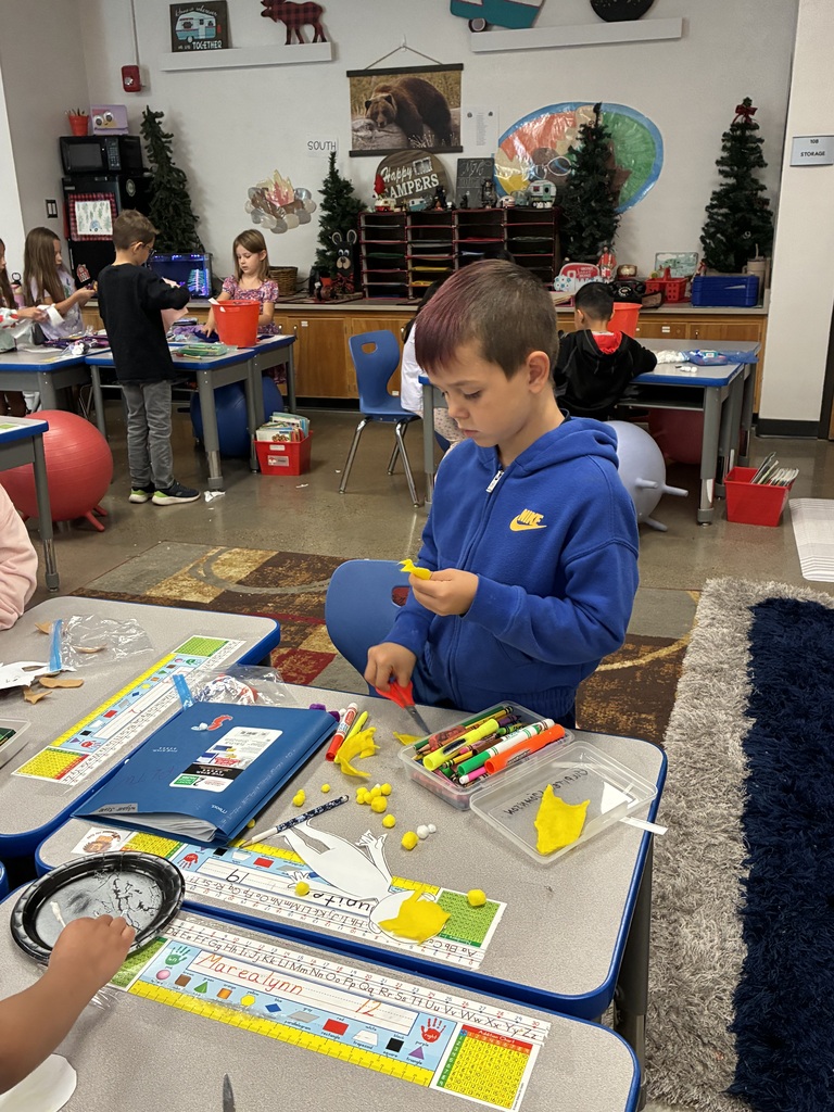 A classroom with students at tables. A boy in a blue hoodie is standing at a table, cutting paper.