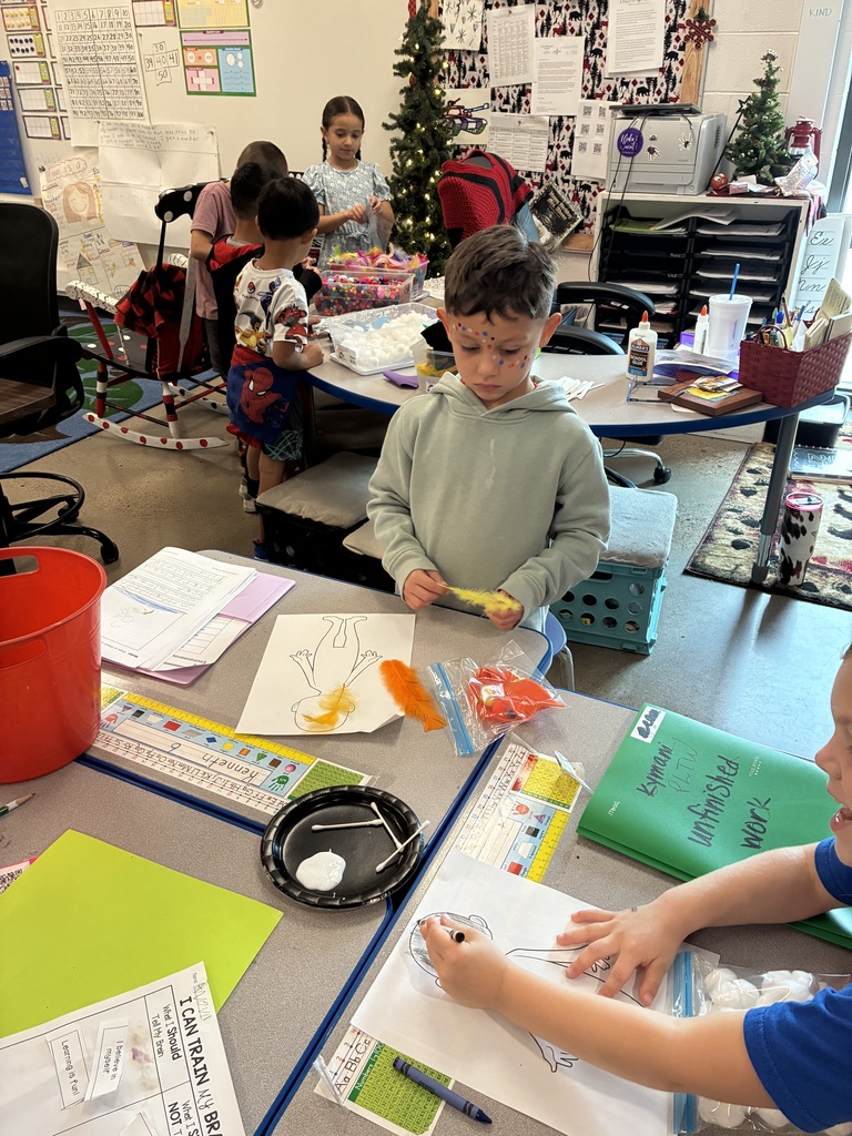 Children in a classroom, one stands at a table with art supplies, another sits at a table drawing.