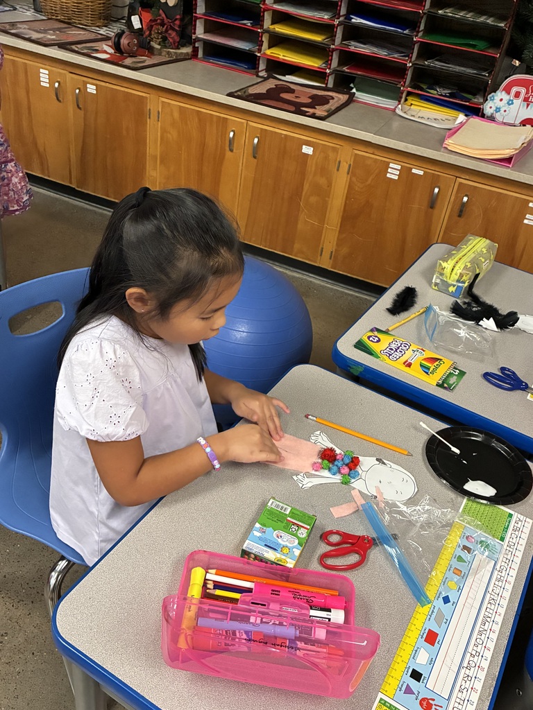 A young girl sits at a table, surrounded by craft supplies, including scissors, a ruler, and a ruler.