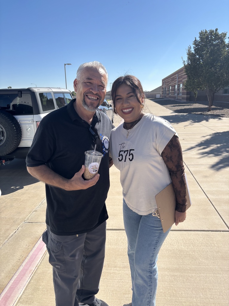 Two people stand outside. A man in black and a woman in white. She holds a folder. Behind them is a parking lot and a building.
