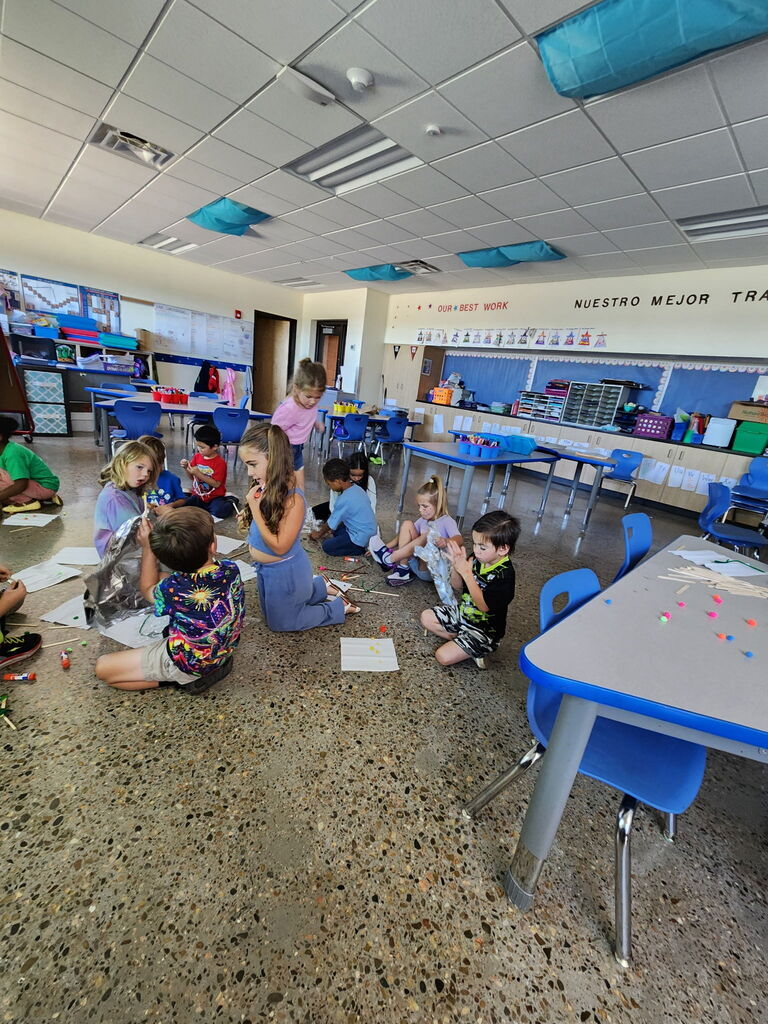 Young children in a classroom, some seated on the floor, others on chairs, engaged in an activity. A table and chairs are in the background.