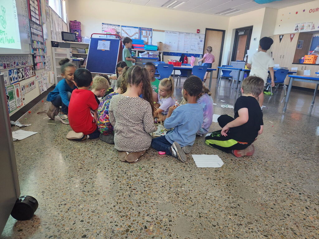 A classroom with children seated in a circle, a woman standing, and others in the background.