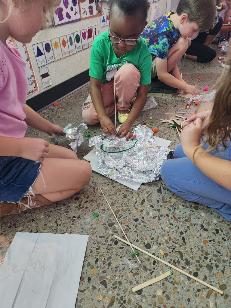 Children seated on the floor, engaged in a craft project with sticks and silver foil, with one child wearing glasses.
