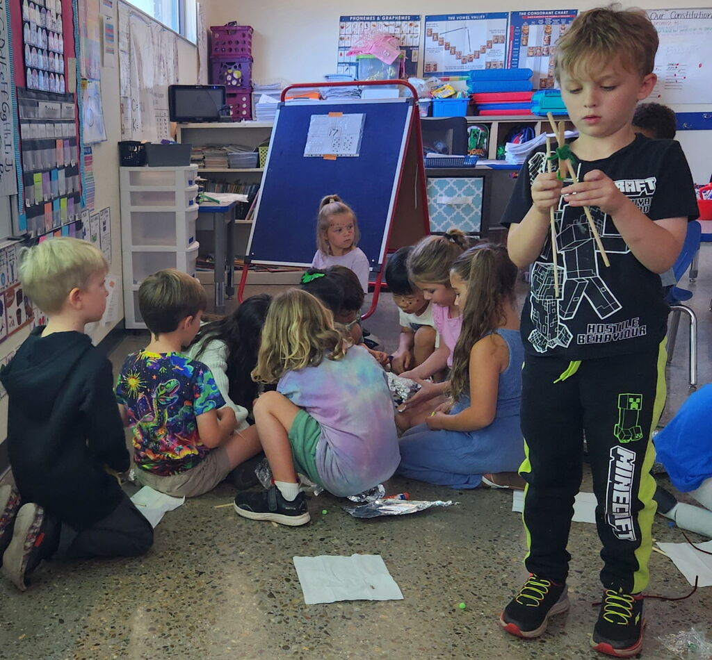 Children seated in a classroom, one stands holding a craft project, with classroom materials in the background.