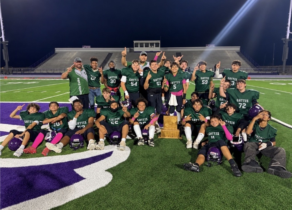 A sports team celebrates on a football field at night, with some members holding a trophy. They wear green jerseys and some have sneakers.
