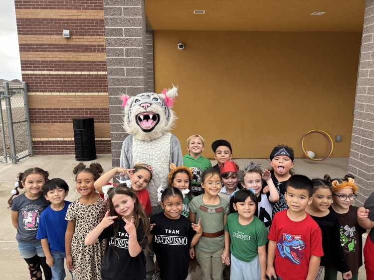 A group of children stand with a mascot in front of a building. The mascot is a white tiger with pink ears.