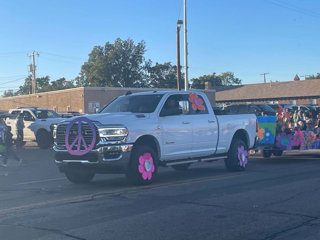 A white pickup truck decorated with a peace sign and pink flowers drives down a street with buildings.